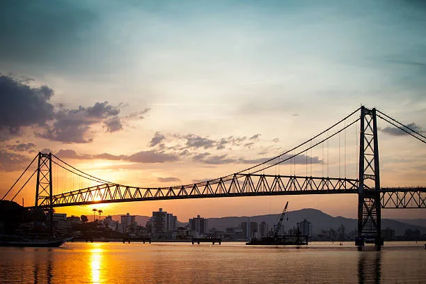 Silhueta da Ponte Hercílio Luz ao pôr do sol sobre a água em Florianópolis.
